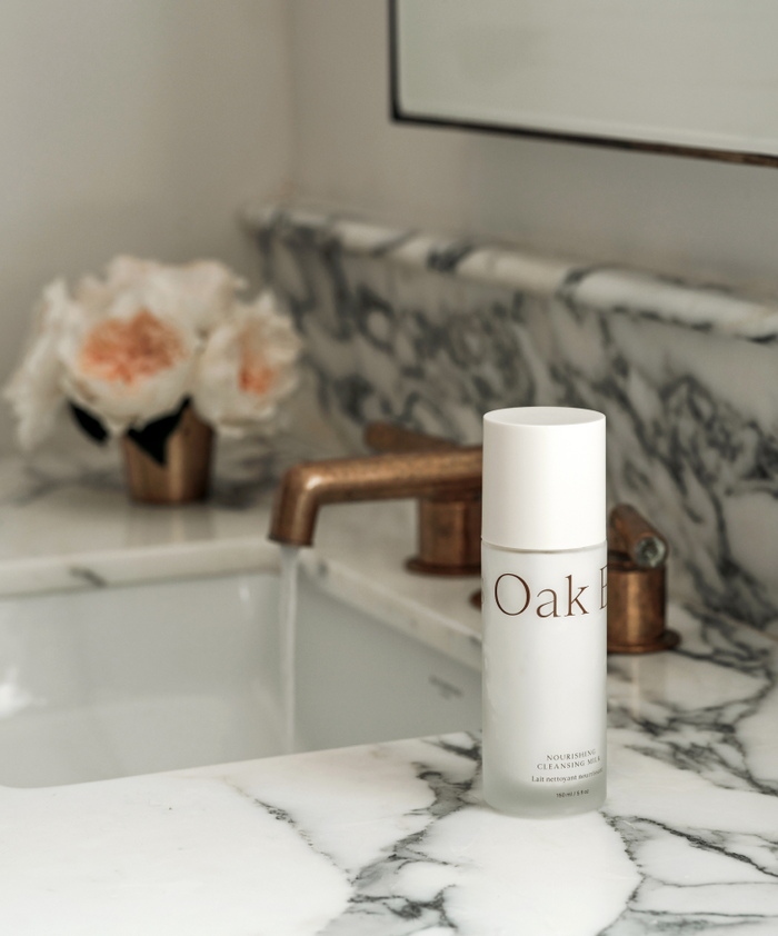 A white bottle labeled Hydrating Double Cleanse Duo, perfect for moisture-rich cleansing, is on a marble bathroom counter by a sink with bronze fixtures. In the background, pale pink flowers sit in a gold vase.