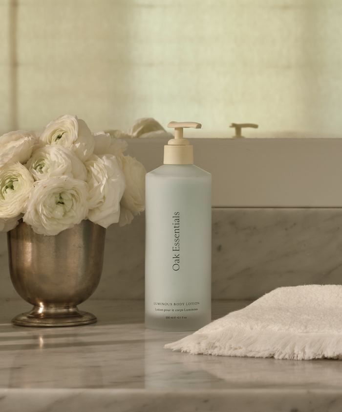 A bottle of Luminous Home and Away Duo sits on a marble countertop beside a silver vase of white flowers and a folded towel, promising silky-soft skin, with a softly lit mirror in the background.