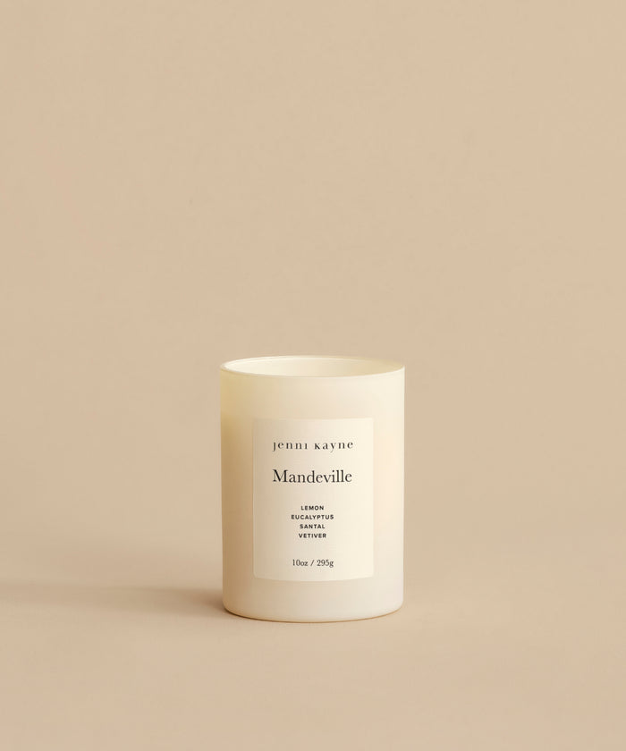A white Mandeville Glass Candle in a minimalist container rests on a beige surface. Its label lists Lemon, Eucalyptus, and Vervain scents and notes that its hand-poured in Los Angeles.