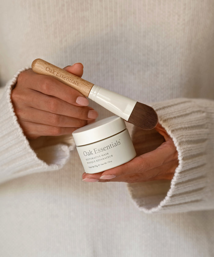 A person in a cream sweater holds a jar of Oak Essentials Moisture Rich Balm and the Mask Brush with a wooden handle, ready for an at-home spa experience.
