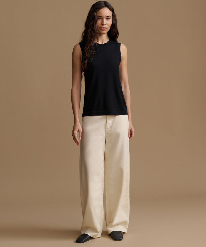 A woman stands against a plain warm beige studio background wearing a sleeveless black top and wide-leg cream pants.