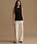 A woman stands against a plain warm beige studio background wearing a sleeveless black top and wide-leg cream pants.
