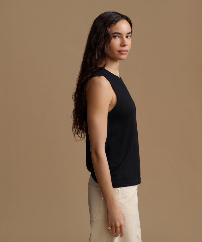 A woman in side profile wearing a black sleeveless top and light beige pants, standing against a solid warm brown studio background.