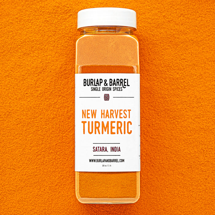 Plastic container of New Harvest Turmeric from Burlap & Barrel with a white lid on a background of loose turmeric powder.