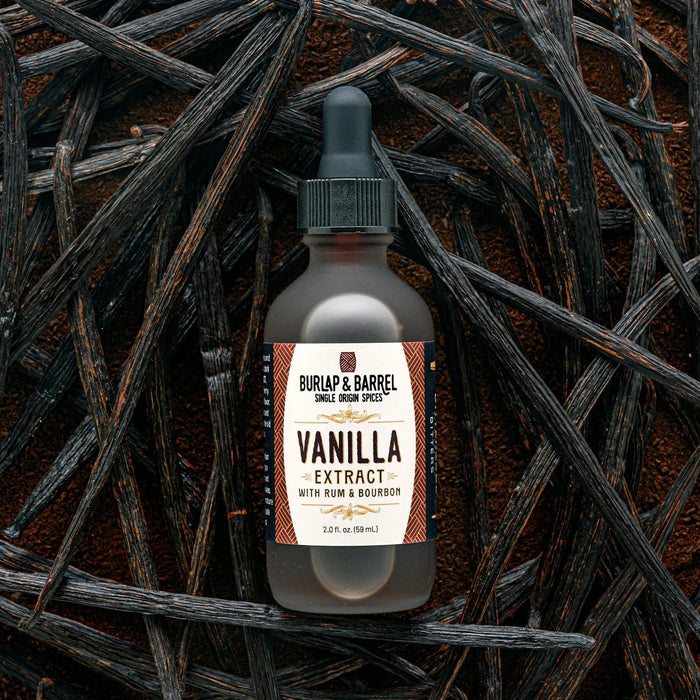 A bottle of Burlap & Barrel Vanilla Extract with Rum & Bourbon, featuring a dark dropper cap and cream label, set against a background of dried vanilla beans.