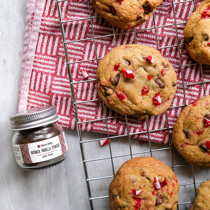 Chocolate chip cookies with red candies on a cooling rack next to a jar of vanilla powder.