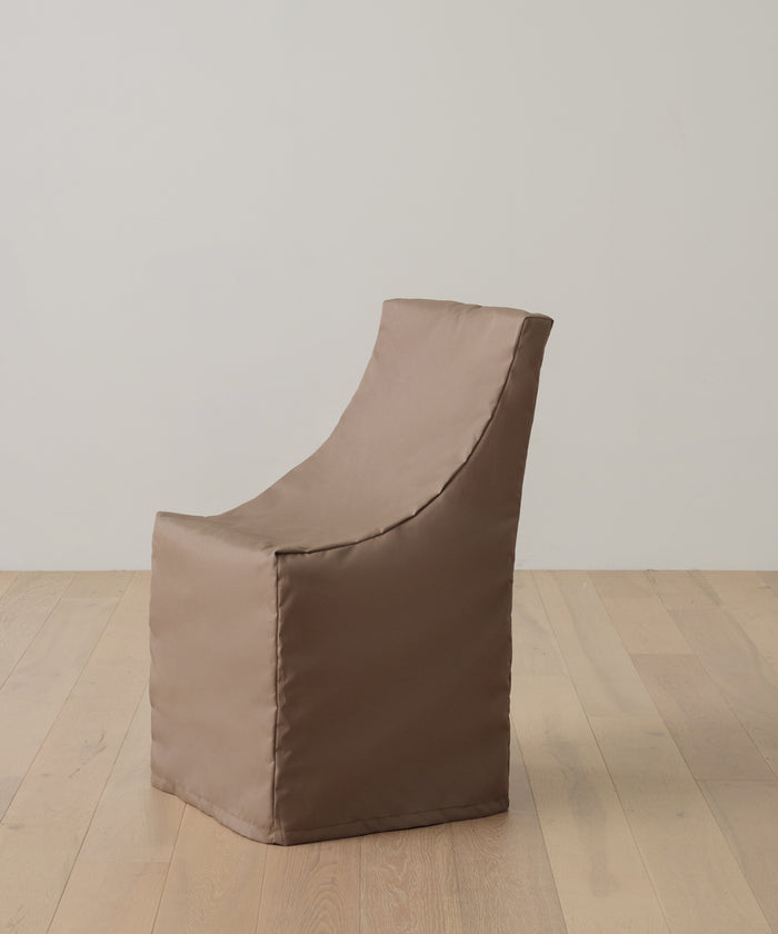 A brown, fabric-covered armchair with a minimalist design sits on a light wooden floor against a plain white wall, shielded by the Outdoor Montecito Dining Chair Cover for UV resistant, long-lasting protection.