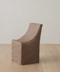 A brown, fabric-covered armchair with a minimalist design sits on a light wooden floor against a plain white wall, shielded by the Outdoor Montecito Dining Chair Cover for UV resistant, long-lasting protection.