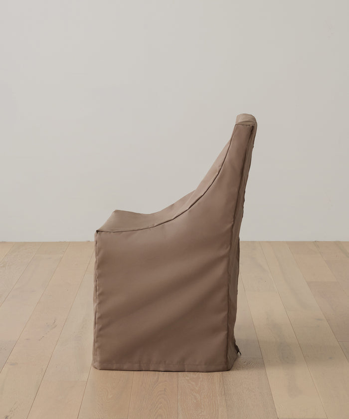The Outdoor Montecito Dining Chair Cover in brown is displayed in profile on a light wooden floor, set against a plain off-white wall.