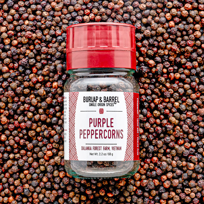 A glass jar of Purple Peppercorns with a red lid, placed on a background of loose purple peppercorns spilling out. The label on the jar reads 'Purple Peppercorns, Single Origin Spices, Burlap & Barrel'.