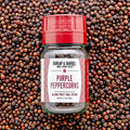 A glass jar of Purple Peppercorns with a red lid, placed on a background of loose purple peppercorns spilling out. The label on the jar reads 'Purple Peppercorns, Single Origin Spices, Burlap & Barrel'.