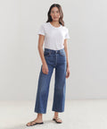 A woman stands indoors against a plain light background, wearing a white short-sleeve t-shirt, black sandals, and high-waisted vintage wide-leg denim Ren Jeans.
