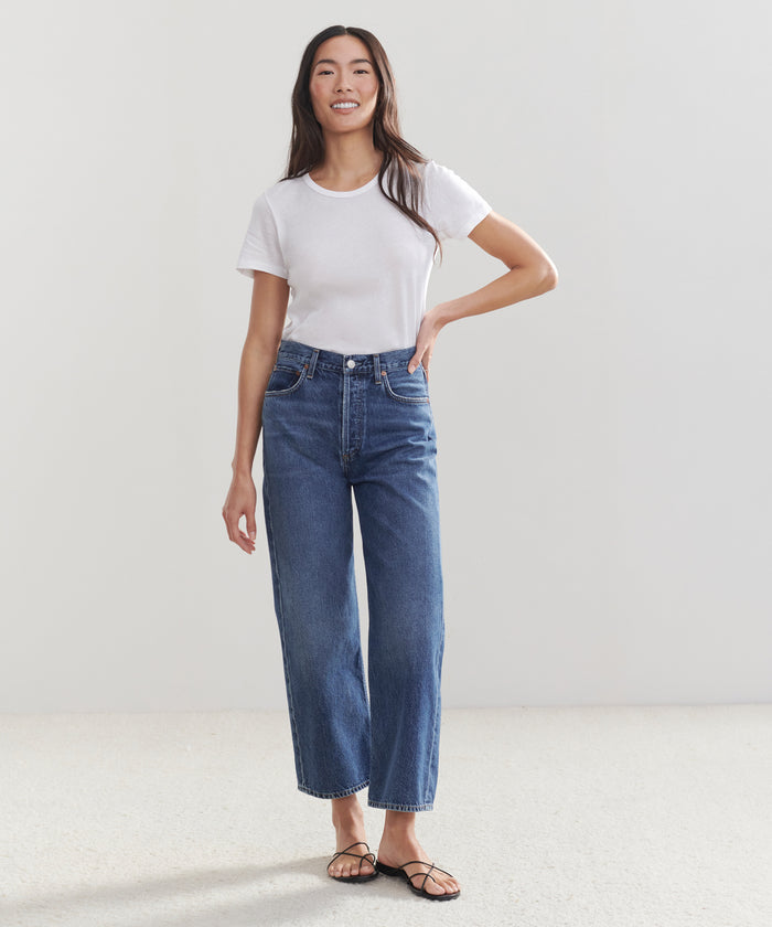 A woman smiles against a plain background, wearing the Ren Jean—high-waisted blue jeans made from sustainable denim—paired with a white t-shirt and black sandals, one hand resting on her hip.