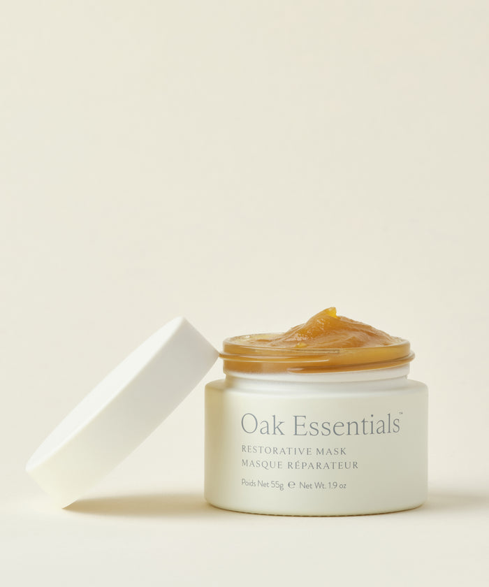An open jar of Restorative Mask with its white lid beside it displays the golden, gel-like mask, enriched with Avocado Oil and organic honey, set against a plain light background.