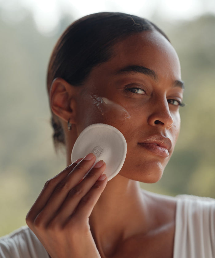 A person with smooth skin uses Reusable Cotton Rounds to gently apply or remove skincare from their cheek, showcasing an eco-friendly cleansing routine against a softly blurred outdoor background.