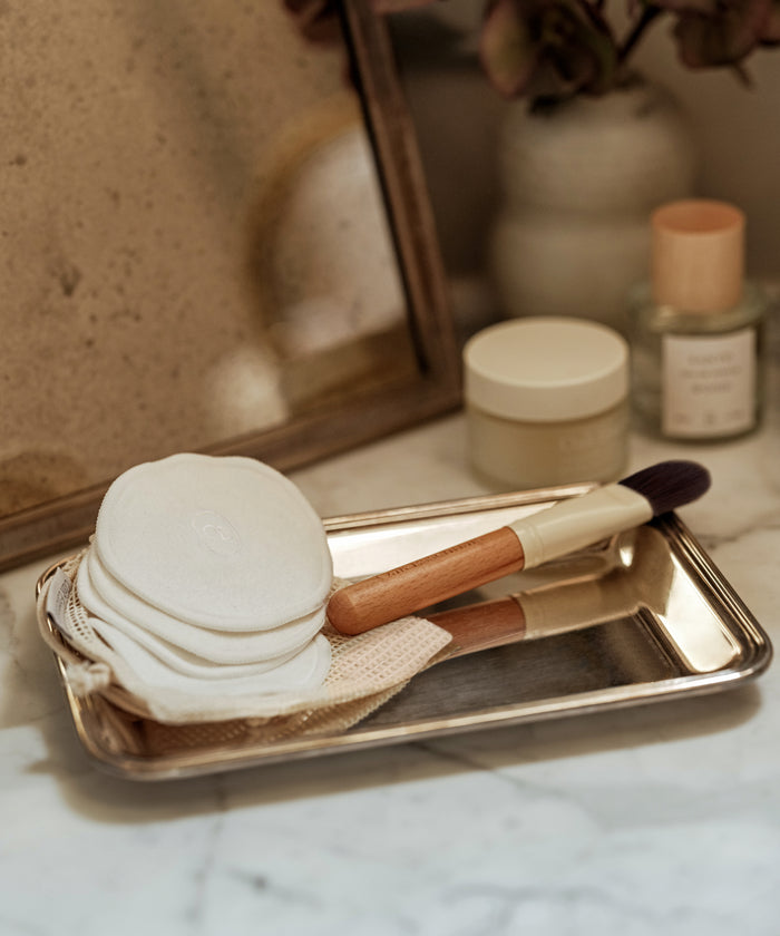 A silver tray on marble holds Reusable Cotton Rounds, a wooden-handled makeup brush, and a washable cotton mesh bag—ideal for an eco-friendly cleansing routine. Cosmetic containers and a framed mirror are in the background.