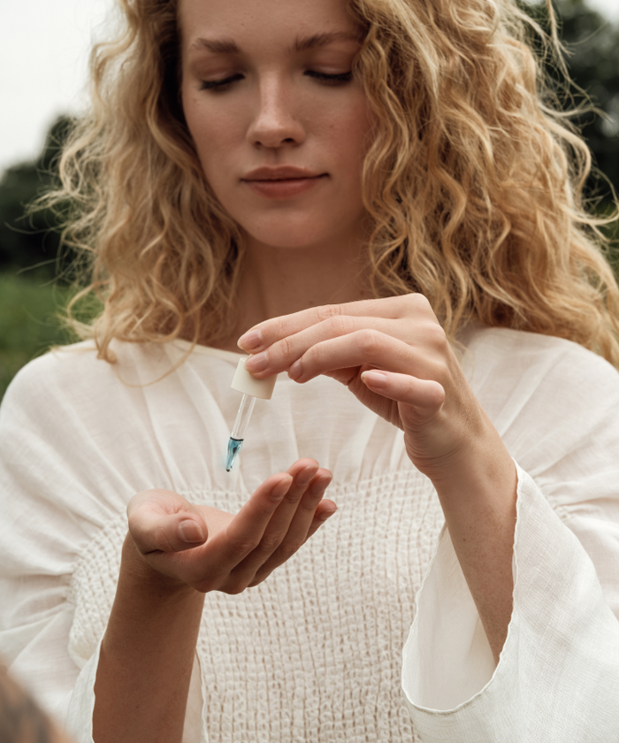 A woman with curly blonde hair in a white blouse dispenses Ritual Oil from a dropper onto her palm outdoors, nourishing her sensitive skin.