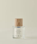 A clear glass bottle of Santo Wood Eau De Parfum with a wooden cap rests on a light beige surface. Its label shows 50 ml (1.7 fl oz). The clean, non-toxic fragrance stands out against the plain, light-colored background.