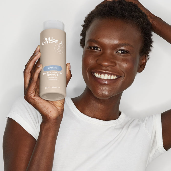 Image of Paul Mitchell model holding Sheer Hydration Shampoo bottle near hear face in a white shirt on a neutral background