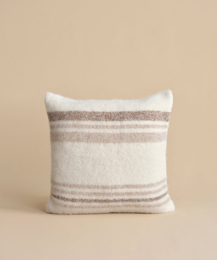 The Sheridan Pillow, a square cushion featuring horizontal beige and light brown stripes on white alpaca blend yarn, is shown upright against a plain beige background.