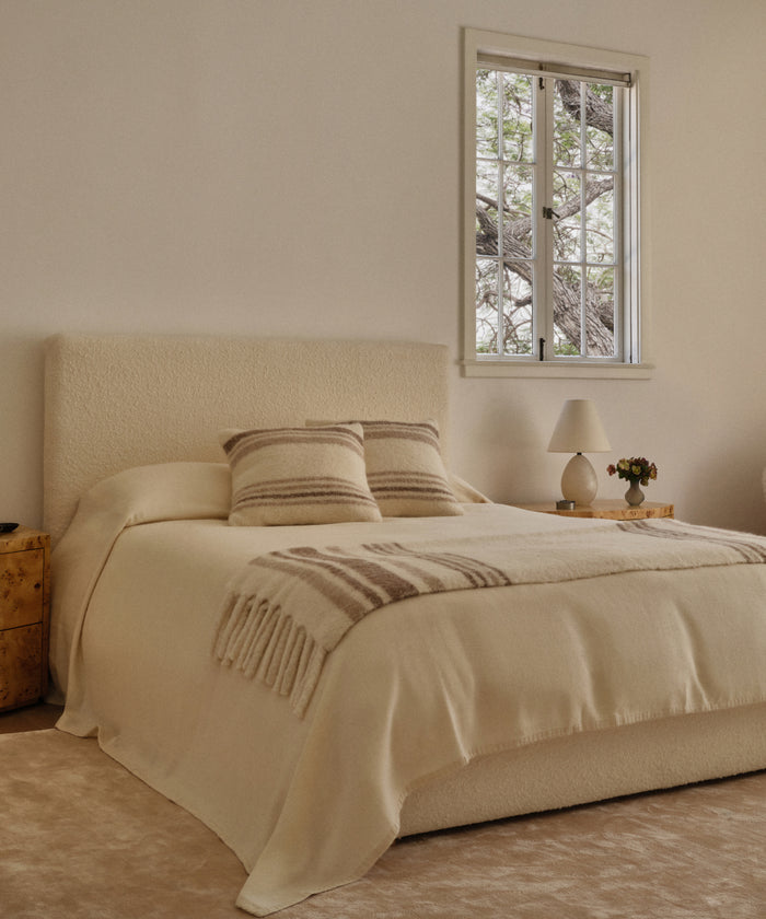A cozy, minimalist bedroom features a cream-colored bed with matching pillows and a Sheridan Pillow. A wooden nightstand holds a lamp and small decor, while a window frames tree branches outside.