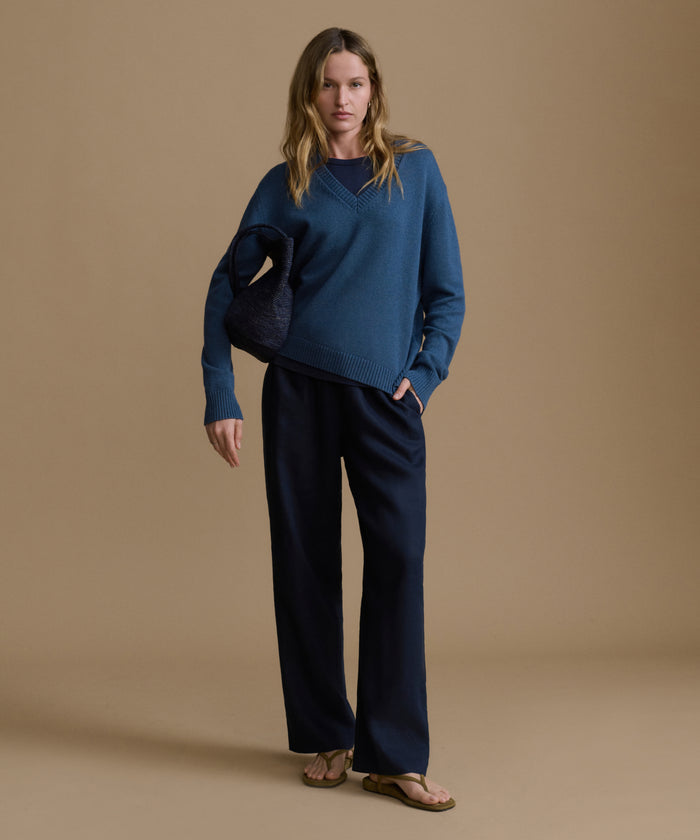 Woman wearing a navy v-neck sweater with blue pants against a brown wall.