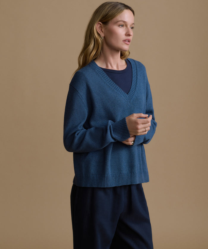 Woman wearing a navy v-neck sweater with blue pants against a brown wall.