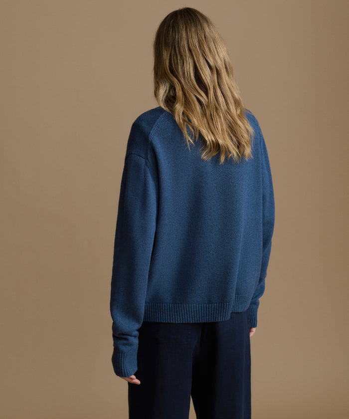 Woman wearing a navy sweater with blue pants against a brown wall.