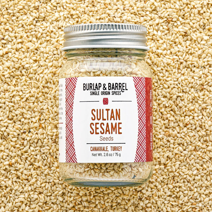 A glass jar of Sultan Sesame seeds by Burlap & Barrel, filled with sesame seeds and topped with a silver metal lid, set against a background of loose sesame seeds. The label features a white background with red patterned borders and brown text.