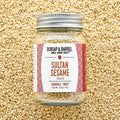 A glass jar of Sultan Sesame seeds by Burlap & Barrel, filled with sesame seeds and topped with a silver metal lid, set against a background of loose sesame seeds. The label features a white background with red patterned borders and brown text.
