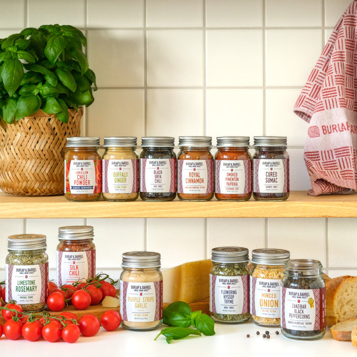 A set of eight glass spice jars from Burlap & Barrel with silver lids, arranged on wooden kitchen shelves alongside fresh ingredients including basil, tomatoes, garlic, onion, and bread against a white tiled wall.