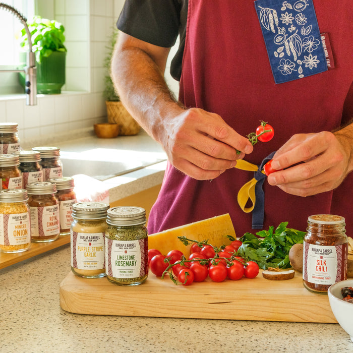 A person wearing a red apron stands in a bright kitchen, holding a small tomato with a ribbon. In front of them is a wooden cutting board with spices, fresh tomatoes, herbs, cheese, and olives, alongside multiple glass spice jars on a countertop.