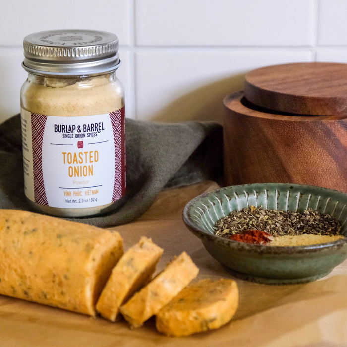 Jar of Burlap & Barrel toasted onion seasoning on a wooden surface with bread and a bowl of spices.