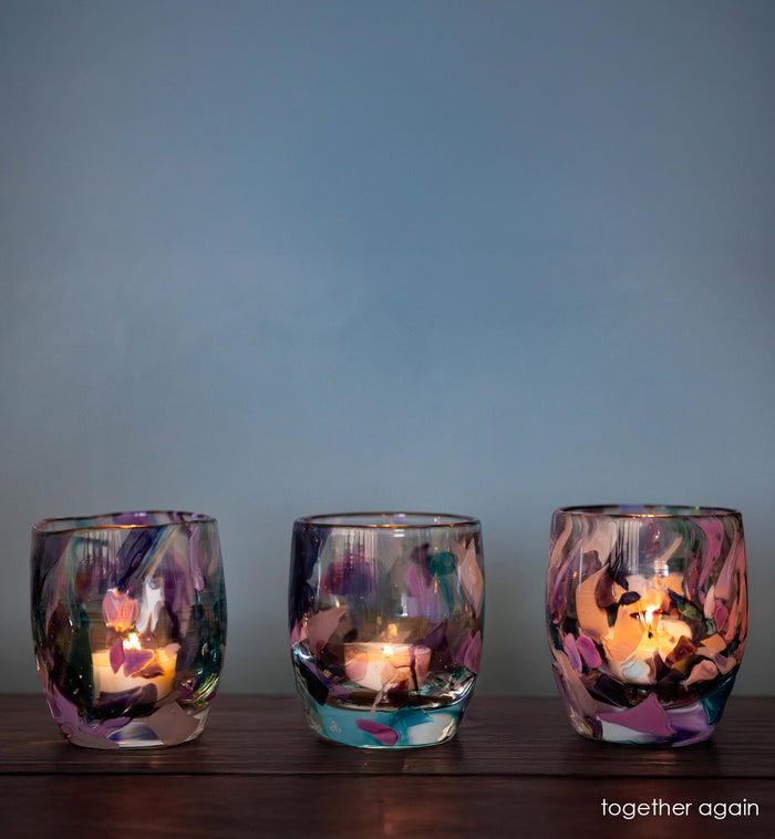 three lit 'together again' multi-color (blue, purple, brown, light blue) hand-blown glass candle holders on wood table.