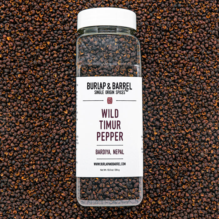 A bottle of Wild Timur Pepper by Burlap & Barrel, with the label showing the product name and origin, amidst loose peppers.