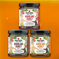 Three jars of Burlap & Barrel Wild Woodland Honey displayed against an orange background, each jar with a label featuring floral designs and the name of the honey variety.
