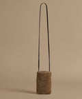 A small cylindrical shoulder bag made of brown textured shearling with a thin black shoulder strap, set against a solid beige background.