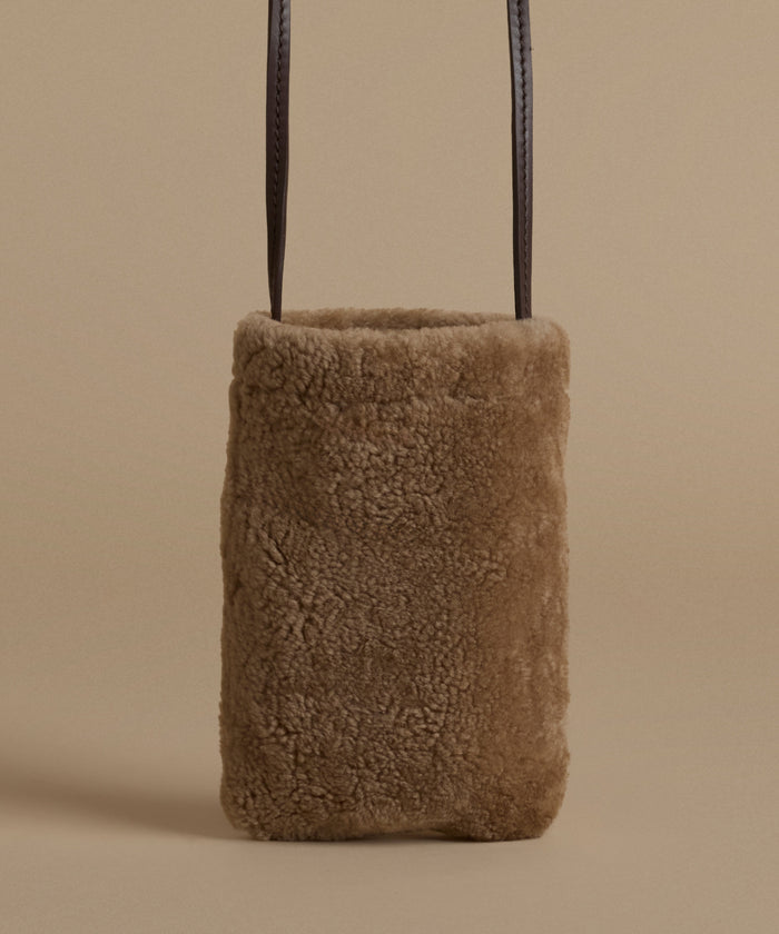 A small rectangular shoulder bag made of soft brown shearling, with dark brown leather shoulder straps, shown against a solid beige background.