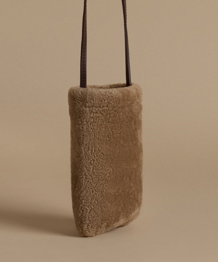 A vertical, rectangular shoulder bag made of soft, textured shearling in a warm taupe-brown color, featuring two slim dark brown leather straps, set against a solid beige background.