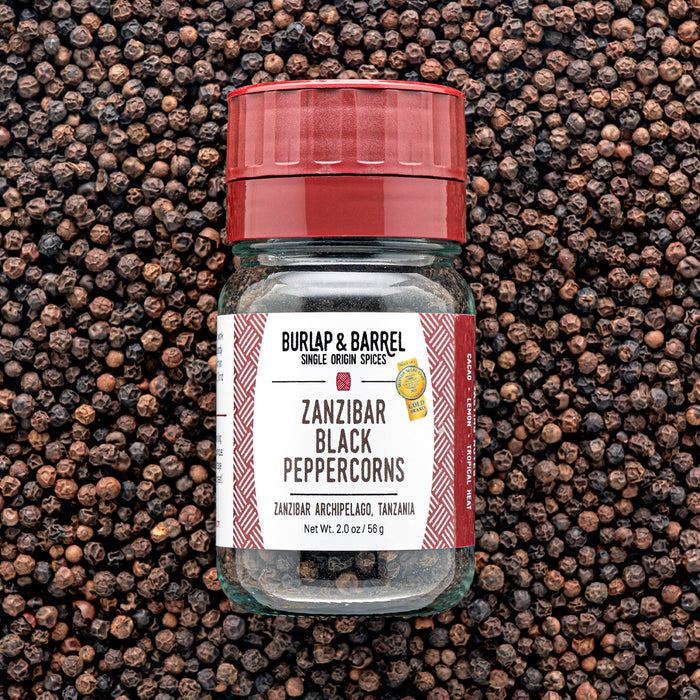 A glass jar of Zanzibar Black Peppercorns from Burlap & Barrel with a red lid, set against a background of loose black peppercorns.