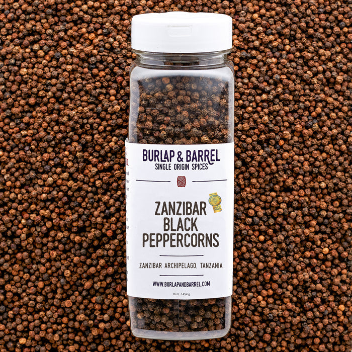 A plastic container of Zanzibar Black Peppercorns from Burlap & Barrel with a white lid, set against a background of loose black peppercorns.